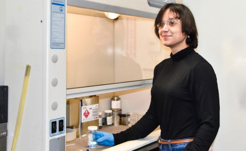 A young women holds a beaker of clear liquid while standing in a lab.